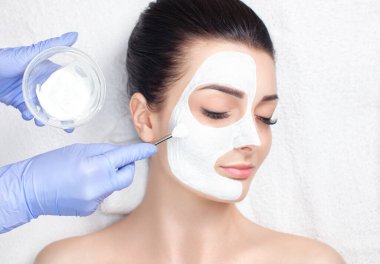 Beautician makes a face clay mask against acne on the face of a woman to rejuvenate the skin. Cosmetology treatment of problem skin on the face and body. clipart