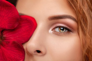 Gözler ve kaşlar kapanır. Güzel makyajlı ve sağlıklı ciltli güzel bir genç kızın portresi. Yüzün yanında kırmızı orkide çiçeği var. Makyaj ve kozmetoloji kavramı.