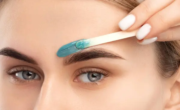 Makyöz, yüz kıllarını aldırma prosedürü uyguluyor. Mavi gözlü güzel bir kız. Kaşlarında daimi makyaj var. Profesyonel makyaj..