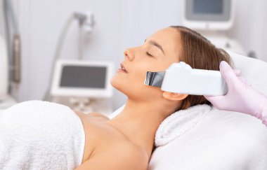 The cosmetologist makes the ultrasound cleaning procedure of the facial skin of a beautiful, young woman in a beauty salon. Cosmetology and professional skin care. clipart