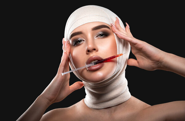 Молодая женщина с бинтом на голове держит шприц для инъекций в губах. Пластическая хирургия, пластика лица.