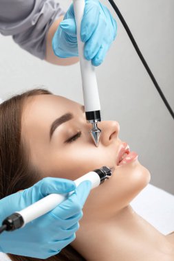 A doctor cosmetologist makes a microcurrent facial therapy to a young woman with a device in a beauty wellness salon.Cosmetology and professional skin care. clipart