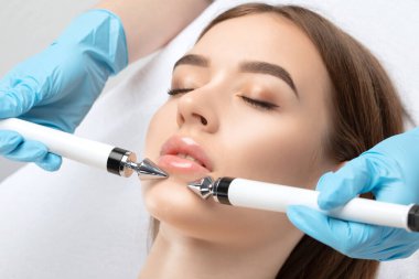 A doctor cosmetologist makes a microcurrent facial therapy to a young woman with a device in a beauty wellness salon.Cosmetology and professional skin care.