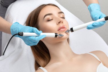 A doctor cosmetologist makes a microcurrent facial therapy to a young woman with a device in a beauty wellness salon.Cosmetology and professional skin care.