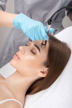 A doctor cosmetologist makes a microcurrent facial therapy to a young woman with a device in a beauty wellness salon.Cosmetology and professional skin care.