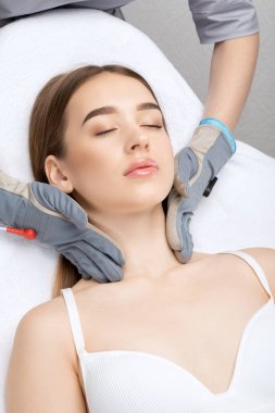 Bir kozmetolog, güzellik salonunda genç ve güzel bir kızın yüzüne ve boynuna mikro akım masaj uygular..