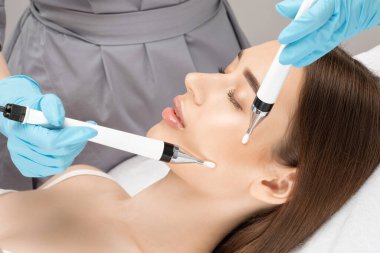 A doctor cosmetologist makes a microcurrent facial therapy to a young woman with a device in a beauty wellness salon.Cosmetology and professional skin care. clipart