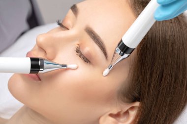 A doctor cosmetologist makes a microcurrent facial therapy to a young woman with a device in a beauty wellness salon.Cosmetology and professional skin care.
