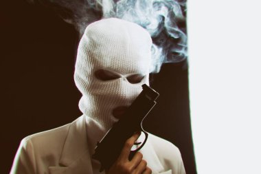 Beyaz kar maskeli bir adam yüzüne yakın bir silahla karanlık bir arka planda dumanlı..