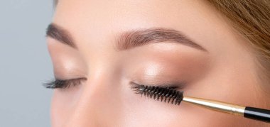 Beautiful young blonde teen girl with long hair, makeup,clean skin. Makeup artist paints eyelashes with mascara in a beauty salon.Eyelash extension procedure.Makeup and cosmetology skin care. clipart