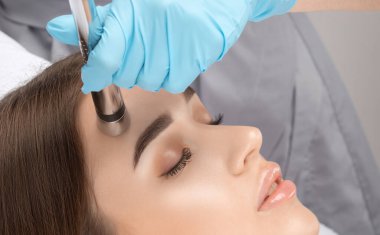 A doctor cosmetologist makes a microcurrent facial therapy to a young woman with a device in a beauty wellness salon.Cosmetology and professional skin care.
