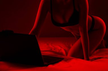 Genç bir webcam mankeni dizüstü bilgisayarın önünde oturuyor. Yatak odasındaki beyaz bir yatakta kırmızı arka planda iletişim kuruyor.