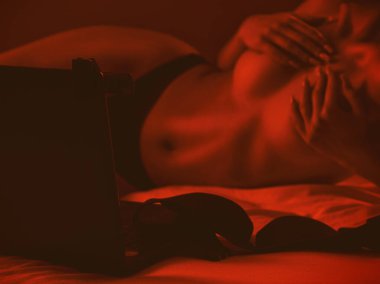 Genç bir webcam mankeni dizüstü bilgisayarın önünde oturuyor. Yatak odasındaki beyaz bir yatakta kırmızı arka planda iletişim kuruyor.
