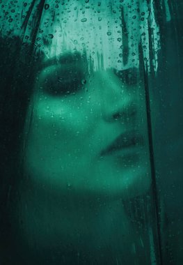 Bir kız pencerenin önünde oturuyor, hava yağmurlu. Pencerede yağmur damlaları görünür.