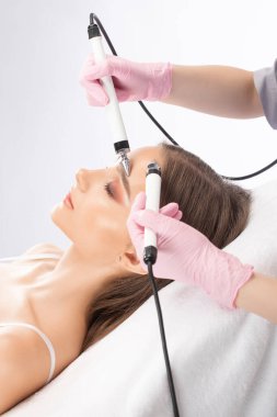 Bir doktor kozmetik uzmanı güzellik salonunda cihazı olan genç bir kadına mikro akım yüz terapisi yapıyor. Kozmetoloji ve profesyonel cilt bakımı.