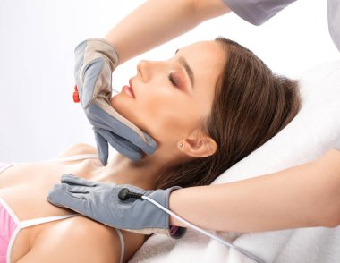 Bir kozmetolog, güzellik salonunda genç ve güzel bir kızın yüzüne ve boynuna mikro akım masaj uygular..