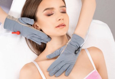 Bir kozmetolog, güzellik salonunda genç ve güzel bir kızın yüzüne ve boynuna mikro akım masaj uygular..