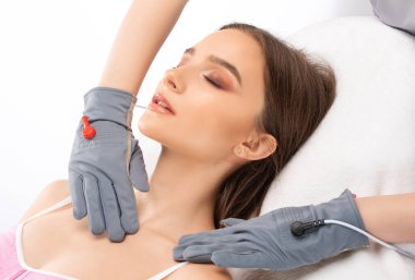 Bir kozmetolog, güzellik salonunda genç ve güzel bir kızın yüzüne ve boynuna mikro akım masaj uygular..