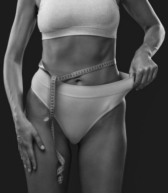 İnce, koyu tenli, bej iç çamaşırlı bir kız koyu arkaplanda bir santimetre ile belini ve kalçasını ölçüyor. Kilo kaybı ve sağlıklı yaşam tarzı kavramı.