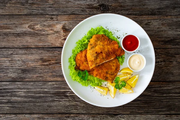 Crispy breaded seared chicken cutlet with lemon slices and fresh vegetables on wooden table