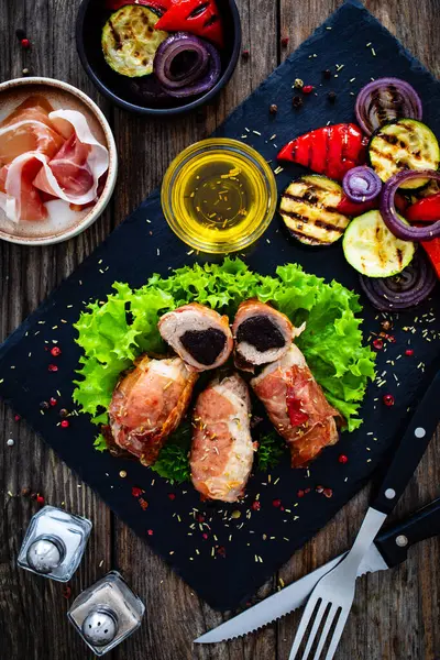 木のテーブルにグリル野菜を添えたハムで包まれた乾燥したプラムで刺された豚肉 — ストック写真