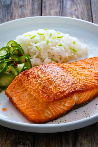Seared salmon steak with boiled white rice and sliced cucumber on wooden table