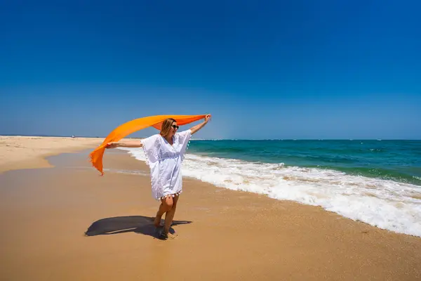stock image Summer vacation. Beautiful young woman in white tunic and sunglasses holding orange scarf fluttering in the wind walking on the sandy beach by the sea with big waves on a beautiful sunny day