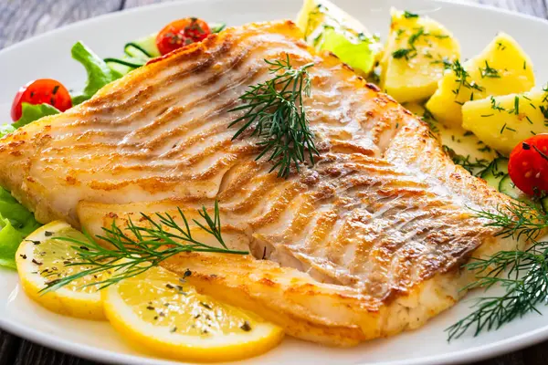 stock image Fried cod fillet, boiled potatoes, fresh vegetables and lemon slices served on white plate on wooden table 