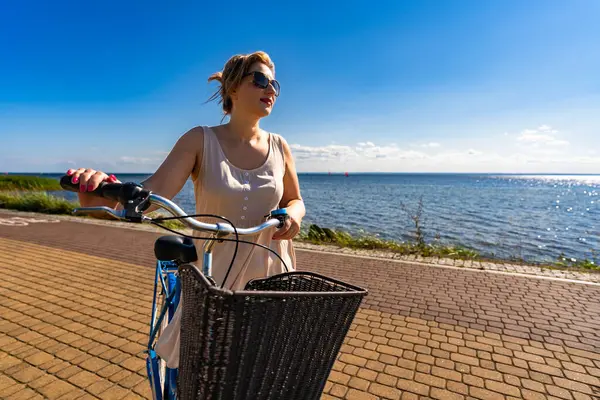 Physical activity at summertime. Portrait of young beautiful woman wearing beige dress and sunglasses standing with city bike on bicycle path on seafront on sunny day. Front view