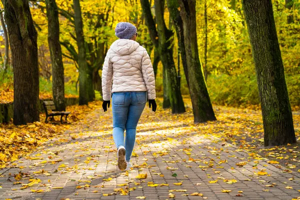 stock image Middle-aged woman wearing jeans, beige jacket and winter hat walking on alley full of yellow fallen leaves in park on autumn day. Back view. Autumnal walk in city park.