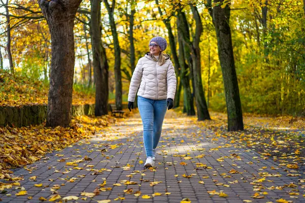 stock image Beautiful middle-aged blonde woman wearing jeans, beige jacket and winter hat and sunglasses walking on alley full of yellow fallen leaves in park on autumn day. Front view. Autumnal walk in city park