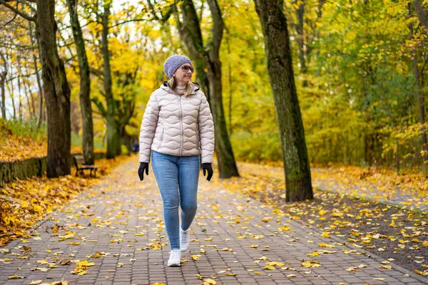 stock image Beautiful middle-aged blonde woman wearing jeans, beige jacket and winter hat and sunglasses walking on alley full of yellow fallen leaves in park on autumn day. Front view. Autumnal walk in city park