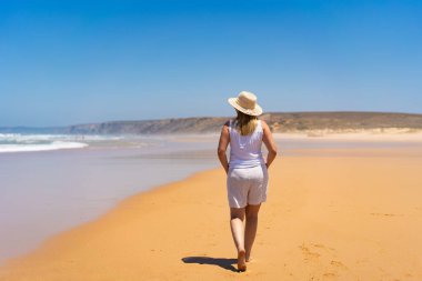 Mid adult beautiful woman wearing light clothes for hot weather, sun hat and sunglasses walking on sandy beach on summer day. Bordeira praia, Portugal. Relax and calm on beach. clipart