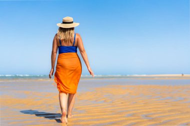 Beautiful middle-aged woman in one-piece blue swimsuit, orange pareo, summer hat and sunglasses walking on sandy beach in summertime. Front view. Armona Island, Portugal. clipart