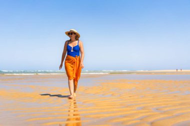 Beautiful middle-aged woman in one-piece blue swimsuit, orange pareo, summer hat and sunglasses walking on sandy beach in summertime. Front view. Armona Island, Portugal. clipart