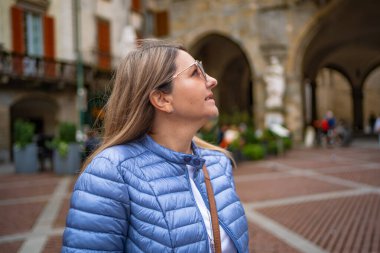 Piazza Vecchia, Bergamo, Italy. Portrait of pretty young blonde woman wearing blue jacket, white blouse and sunglasses standing against old beautiful building on main square in old town on autumn day. clipart