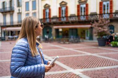 Piazza Vecchia, Bergamo, Italy. Beautiful middle-aged woman wearing blue jacket, white blouse, handbag and sunglasses visiting old town, holding map in her hand on autumn day. Side view. clipart