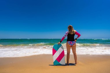 Beautiful young blonde woman in long sleeve swimsuit standing with multicolored bodyboard on sandy beach by ocean shore on sunny day .Armona praia, Algarve, Portugal. Water sports in summertime. clipart