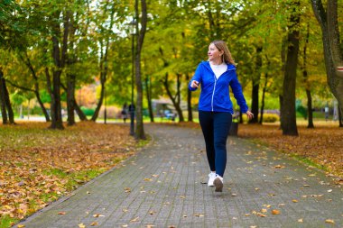 Beautiful mature woman in casual clothing practicing nordic walking in forest full of yellow fallen leaves on autumn day. Front view. Autumnal training of nordic walking. clipart