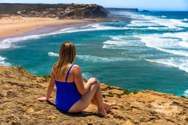 Beautiful mature woman in swimsuit sitting and sunbathing on cliff near sunny beach Monte Clerigo, Algarve, Portugal. Back view clipart