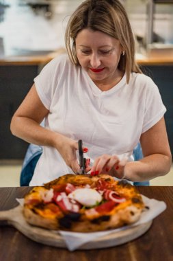 Beautiful mature woman cutting pizza with cured ham and burrata on wooden board into pieces clipart