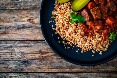 Beef stew with buckwheat groats and dill pickles served on wooden table clipart