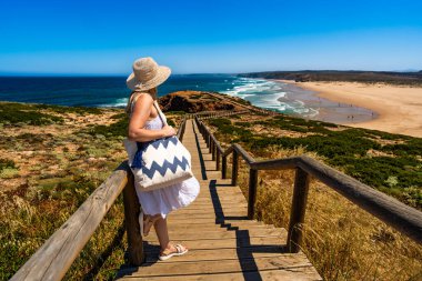 Mature woman tourist admiring picturesque view to cliff, ocean and sandy beach standing on wooden promenade in summertime. Side view. Bordeira beach panorama in Portugal clipart