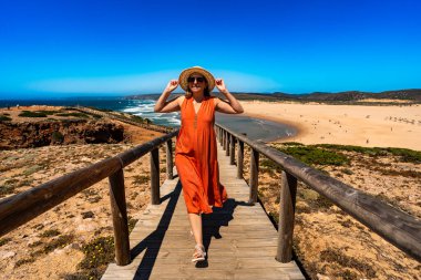 Sightseeing of Algarve coast in Portugal. Happy middle-aged woman tourist enjoying vacations over ocean and beautiful sandy beach - Bordeira. Front view clipart