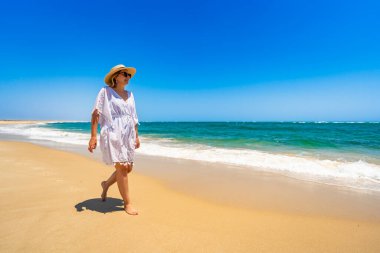 Beautiful mature woman walking on sunny sandy paradise beach on Armona island in Portugal. Travel destination - Armona beach. Side view clipart