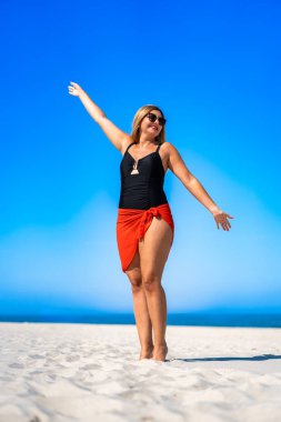 Portrait of beautiful middle aged woman in swimsuit and pareo standing and exposing to sun on white sandy beach in summertime. Model in beautiful pose on beach. Front view clipart