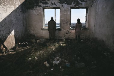 İki kişi, gaz maskeli bir adam ve bir kız, terk edilmiş bir evin içinde, pencerenin yanında duruyorlar ve ileriye bakıyorlar, kıyamete.