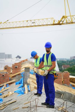 Two construction workers wearing safety vests standing near building materials outdoors