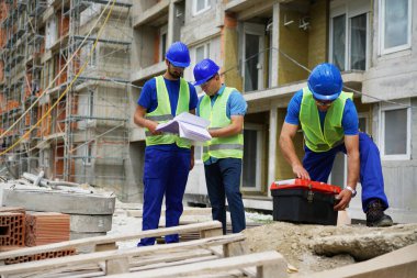Three construction workers wearing safety vests and helmets reviewing blueprints outdoors