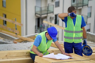 Two construction workers wearing safety vests reviewing blueprints outdoors